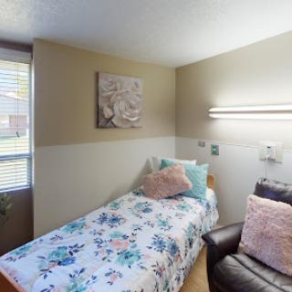 A cozy bedroom featuring a single bed with floral bedding, a comfortable chair, and soft lighting.
