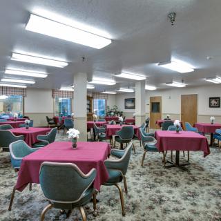 A spacious dining area with multiple tables covered in red tablecloths and surrounded by chairs, illuminated by fluorescent lighting.