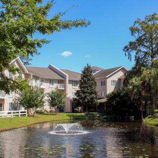 A serene residential building with a pond and fountain in the foreground, surrounded by trees under a clear blue sky.