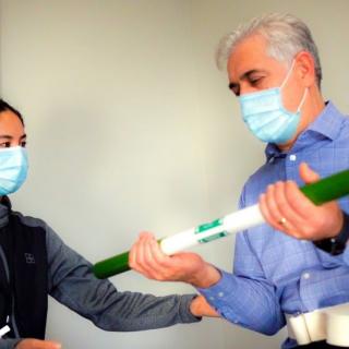 Two individuals wearing masks engage in an exercise or physical therapy session with a resistance bar.