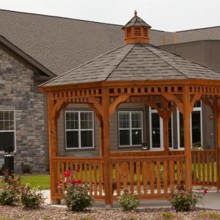 A wooden gazebo stands in front of a house, surrounded by landscaping.