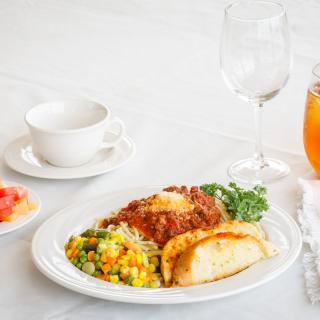 A well-presented dinner plate with lasagna, mixed vegetables, and garlic bread, accompanied by a cup and glasses on a elegant table setting.