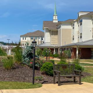 A well-maintained garden with a bench and pathway in front of a residential building with a church steeple in the background under a clear blue sky.