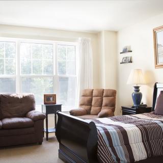 A cozy bedroom with a large window allowing plenty of natural light, featuring a bed, armchair, and sofa.