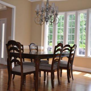 A cozy dining room with a wooden table and chairs, illuminated by a chandelier and natural light from large windows.