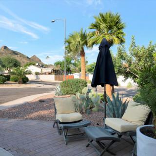 A patio with comfortable outdoor seating and an umbrella, surrounded by palm trees and desert plants in a residential neighborhood.