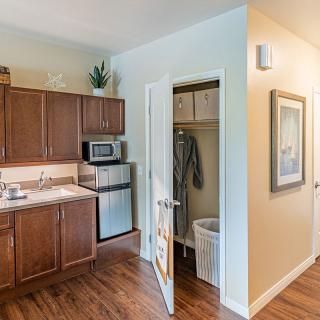 A cozy apartment kitchen with wooden cabinets, a microwave, and a small refrigerator next to an open closet door.