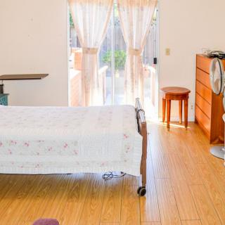 A well-lit bedroom with wooden flooring and simple furniture, including a bed, dresser, and stool.