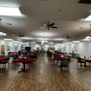 A spacious dining hall with tables and chairs, featuring a wooden floor, ceiling fans, and overhead lighting.