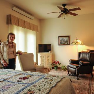 A cozy bedroom with a woman standing near an armchair, featuring a bed, TV, and warm lighting.