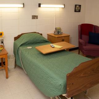 A simple patient room featuring a bed with green bedding, a bedside table, and a comfortable chair.