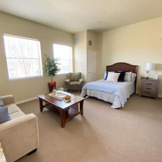 A well-lit bedroom with a bed, sofa, chairs, and a coffee table on a carpeted floor.