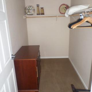 A small closet with a wooden dresser, hanging rod with hangers, and a shelf containing decorative items.