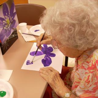 An elderly person painting a purple flower at a table.