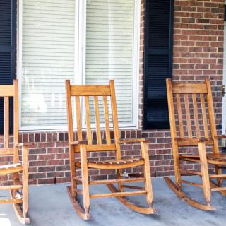 Three wooden rocking chairs sit on a porch in front of a brick wall and windows with shutters.