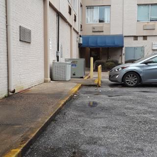 A view of a parking area next to a building, featuring a gray car and an air conditioning unit.