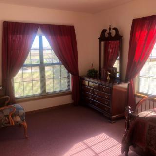 A cozy bedroom featuring a window with red curtains, a wooden dresser with a mirror, and a comfortable chair.