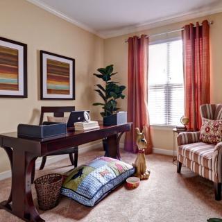 A cozy home office with a dark wooden desk, striped chair, colorful artwork, and a large window with red curtains.