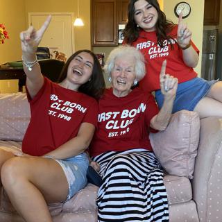 Three women from different generations sit together on a couch, all wearing matching 'First Born Club' shirts and smiling for the camera.