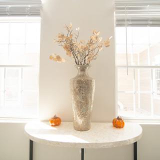 A minimalist white table with a decorative vase of dried flowers and two small pumpkins.