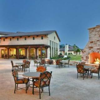 An inviting outdoor patio area with a warm fireplace, surrounded by tables and chairs, set against the backdrop of a large building in the evening.