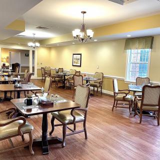 A spacious dining room with neatly arranged tables and chairs, featuring a warm wooden floor and ample lighting.