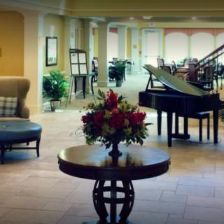 An elegant hotel lobby featuring a grand piano and a floral arrangement on a central table, with comfortable seating and large windows in the background.