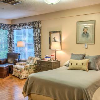 A cozy bedroom with a bed, armchair, and warm lighting from lamps near large windows.