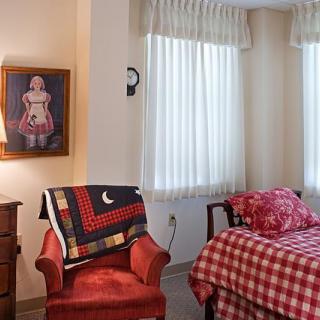 A cozy bedroom with a red-patterned bedspread, an upholstered chair draped with a quilt, and artwork on the wall next to two curtained windows.