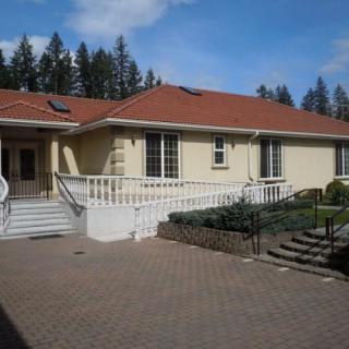 A single-story residential house with a red roof, featuring a paved driveway and surrounded by trees.
