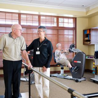 An elderly man receives assistance from a therapist in a rehabilitation center while another senior engages with exercise equipment.