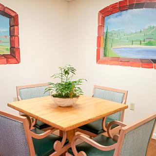 A cozy dining nook with a wooden table, surrounded by four chairs and adorned with an indoor plant. The walls feature murals of scenic landscapes.
