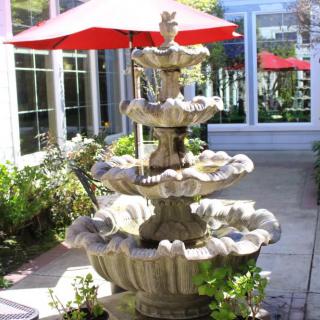A multi-tiered stone fountain with flowing water situated on a sunlit patio next to a building, surrounded by greenery and under the shade of red umbrellas.