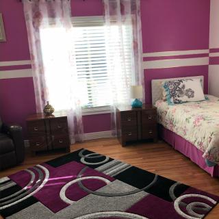 A cozy bedroom features purple walls, a decorative rug, and a comfortable bed near a window with sheer curtains.