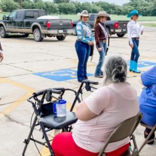 Women in cowboy hats stand in a parking lot while people sit nearby, observing the event.
