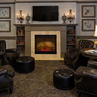 A cozy living room with a fireplace surrounded by leather chairs and ottomans, topped with a television and adorned with decorative sconces and framed art.