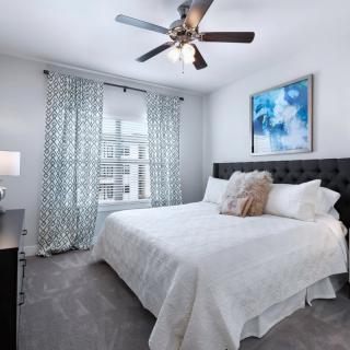 A modern bedroom with a large bed, white bedding, and a ceiling fan. The room features patterned curtains, a piece of abstract artwork above the bed, and a lamp on the nightstand.