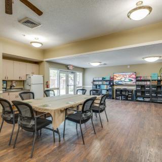 A community room featuring a table and chairs, a kitchen area with a refrigerator, bookshelves, and a television.