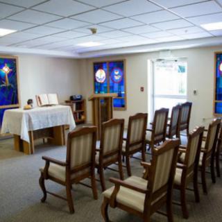 A small chapel with rows of chairs and an altar, featuring stained glass windows.