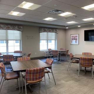 An empty cafeteria with several tables and chairs, featuring windows and a television on the wall.