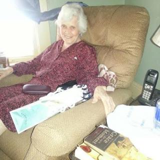 An elderly person sitting comfortably in a recliner surrounded by books and a remote control.