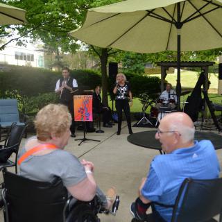 An outdoor event with musicians performing under umbrellas, while an audience watches from their seats.