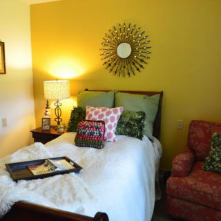 A cozy bedroom with colorful pillows, a decorative mirror, and a comfortable armchair.