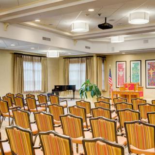A spacious meeting room with rows of chairs facing a stage, featuring a piano and an American flag.