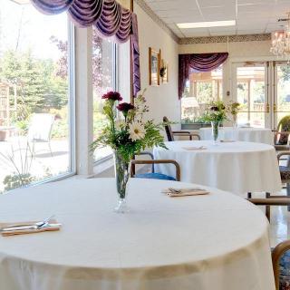A bright dining area with round tables adorned with white tablecloths and floral centerpieces, complemented by large windows and decorative curtains.