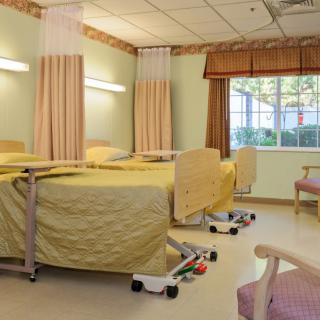 A hospital room with two beds, curtains, and chairs, featuring a window with natural light.
