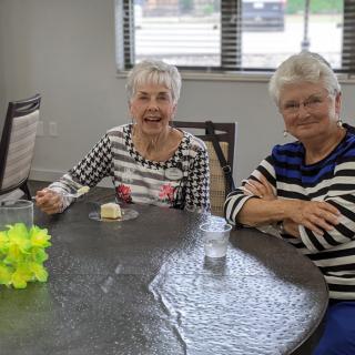 Two elderly women smiling and seated at a table indoors.
