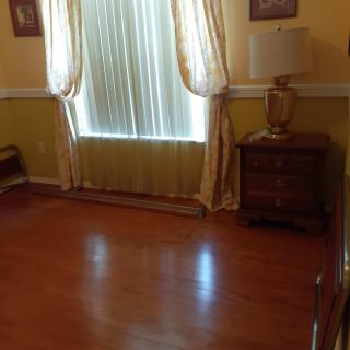 An empty room with hardwood floors, a window with curtains, and a dresser with a lamp on top.