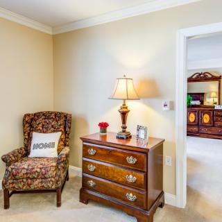 A cozy corner of a living room featuring an armchair, a wooden dresser with a lamp, and a view into another room.