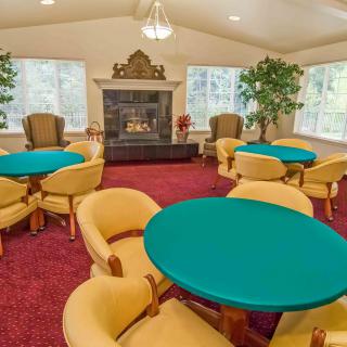 A spacious lounge area featuring multiple round tables and chairs, centered around a fireplace with large windows and indoor plants.